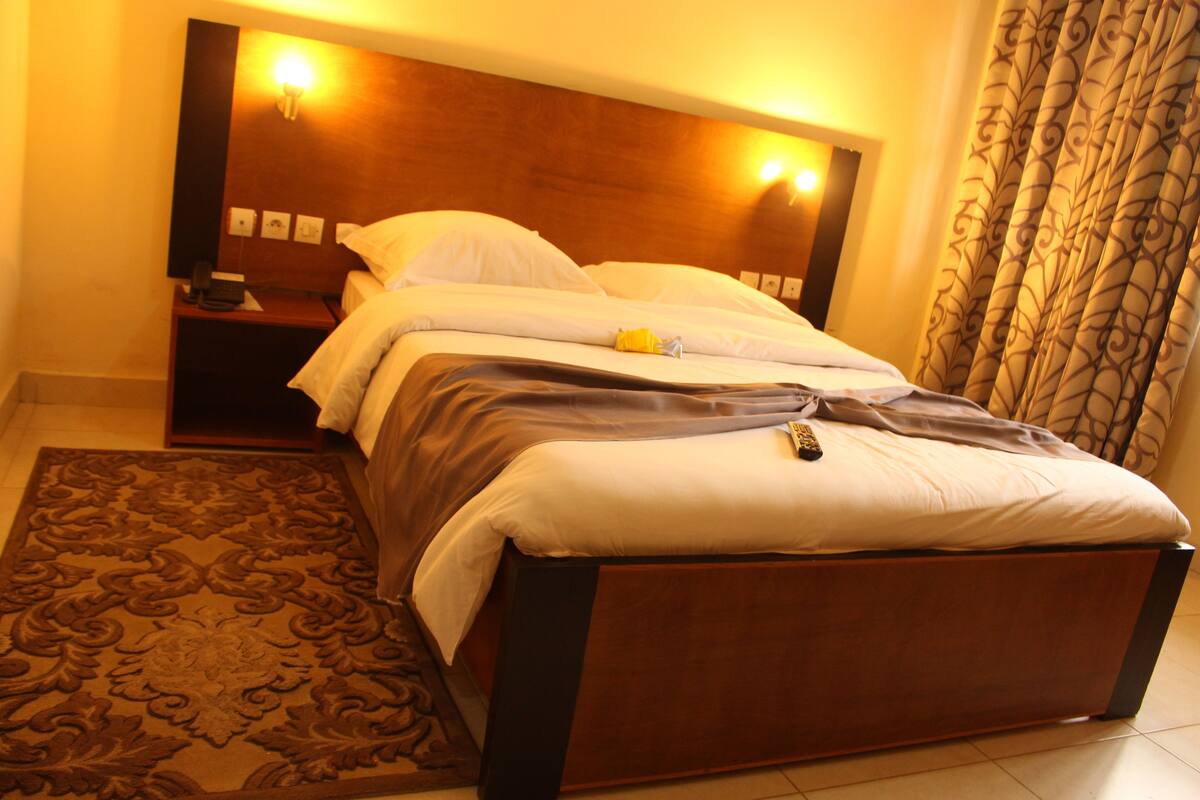 A hotel room with a wooden headboard, a bed with white linens, a bedside table with a lamp, and a patterned rug.