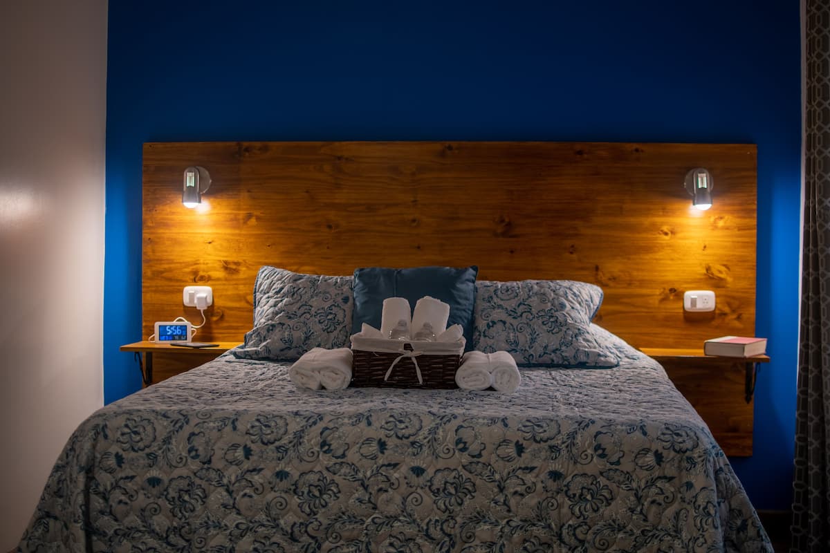 Una cama bien tendida con una colcha de estampado floral, una cesta de toallas y dos lámparas de noche sobre mesitas de madera.