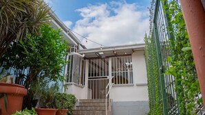 Un espace extérieur abrité, avec des marches menant à une entrée fermée par un portail, entouré de plantes en pot et sous un ciel dégagé.