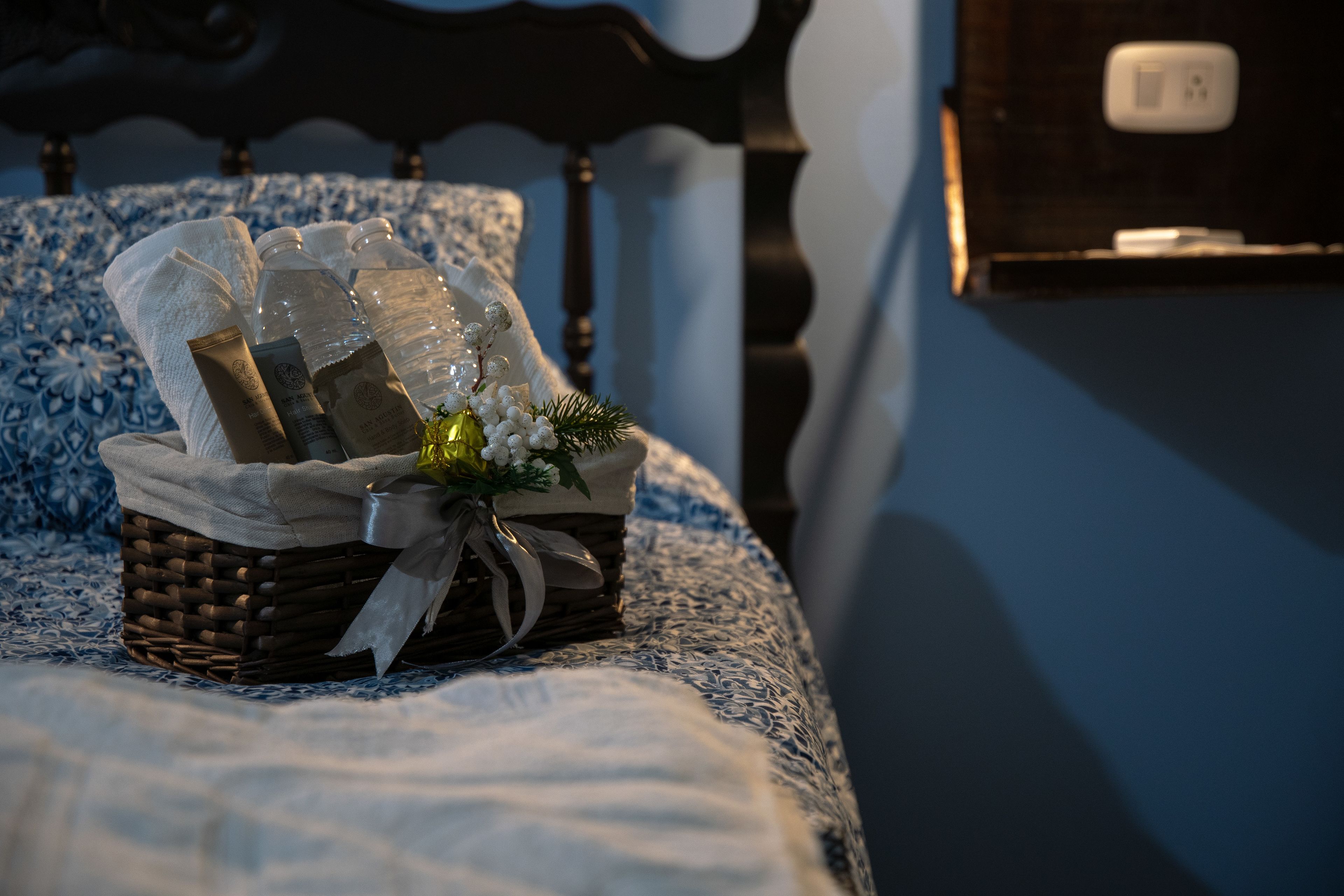 A bed with a blue and white patterned bedspread, a wicker basket with toiletries, and a small bouquet of flowers.