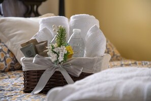 Rottingista punottu kori, jossa on hygieniatarvikkeita, vesipullo ja pieni kasvi, on sijoitettu sängylle, jonka päällä on valkoisia pyyhkeitä.