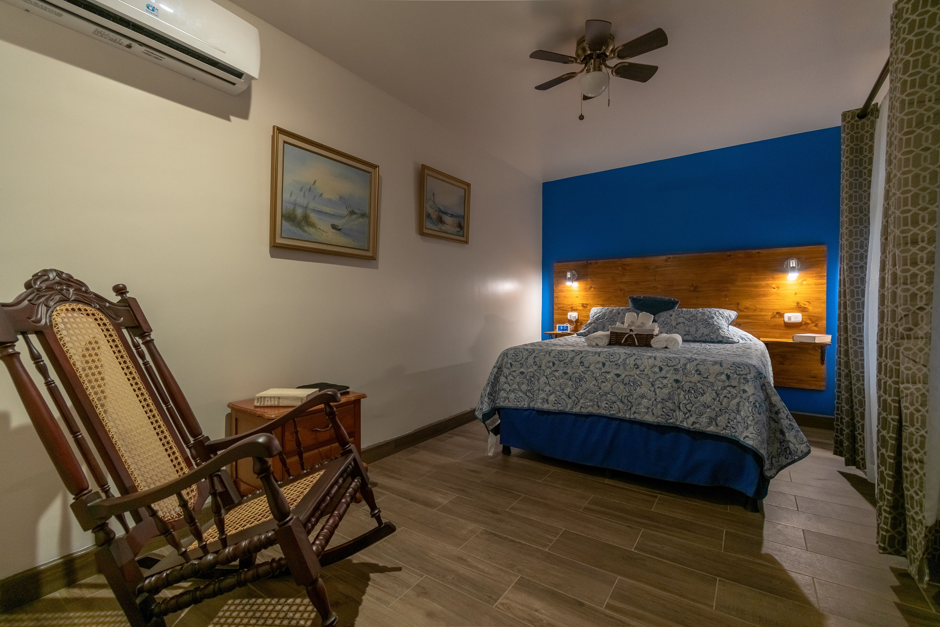 A bedroom with a wooden headboard, a bed with a blue and white patterned bedspread, two wall-mounted lamps, and a ceiling fan.