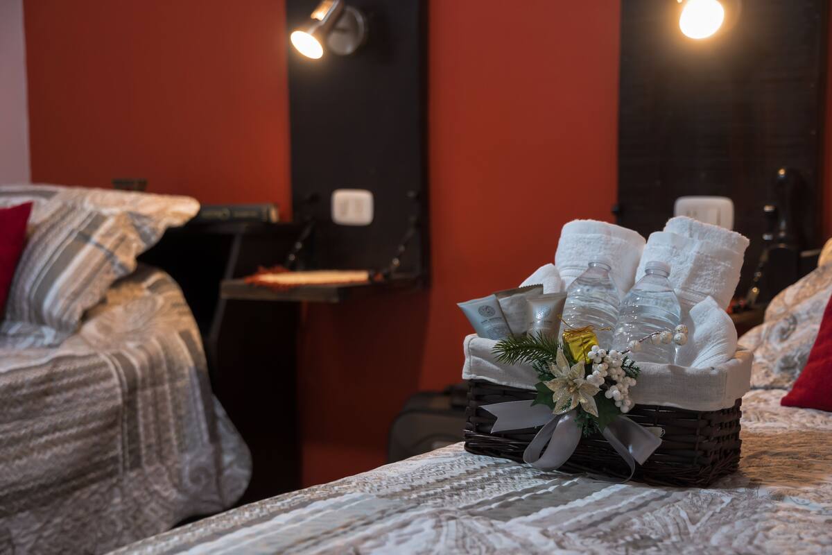 Una habitación de hotel con una cama ordenada, una mesita de noche con lámpara y una cesta decorativa con artículos de aseo.