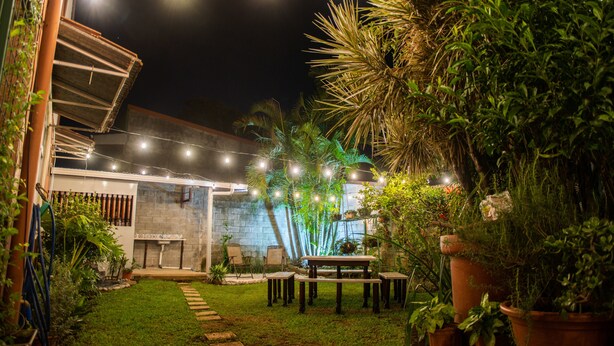Un espace extérieur bien éclairé, aménagé en patio, avec des guirlandes lumineuses, une petite table et des chaises, entouré de plantes en pot et d’une pelouse.
