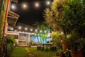 Un espace extérieur bien éclairé, aménagé en patio, avec des guirlandes lumineuses, une petite table et des chaises, entouré de plantes en pot et d’une pelouse.