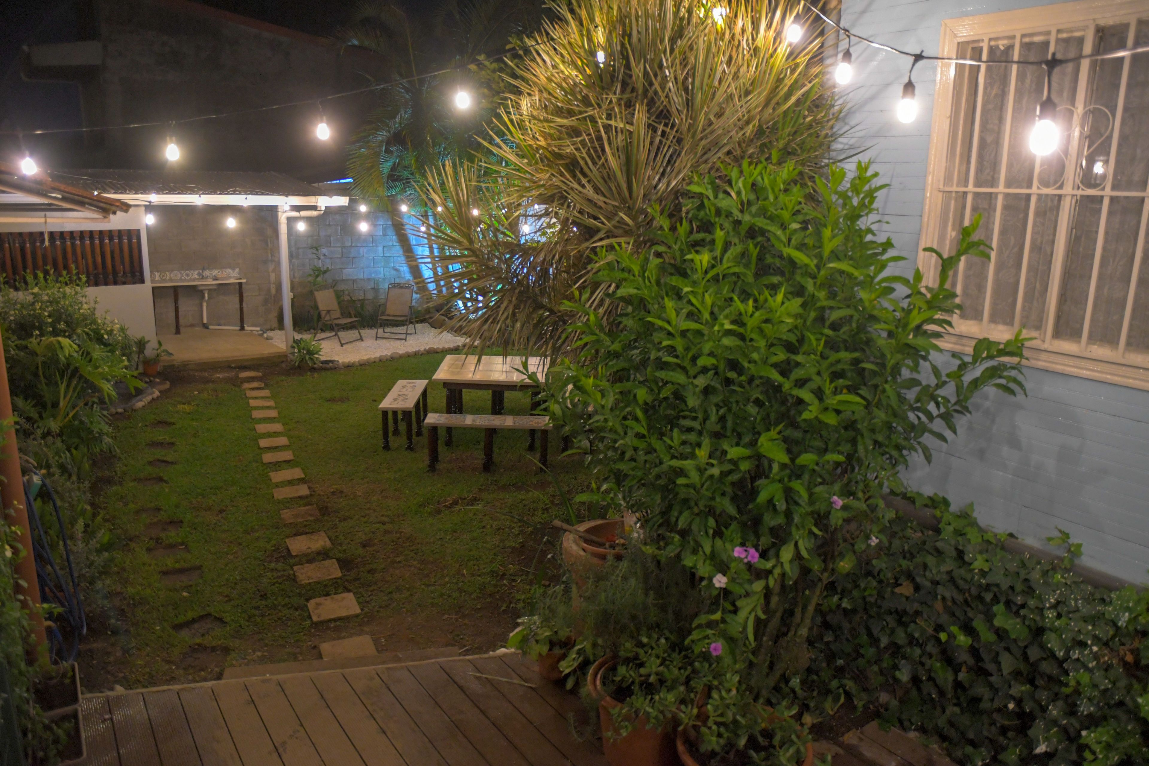A patio area with a wooden deck, a table, and chairs. There are potted plants and a string of lights.