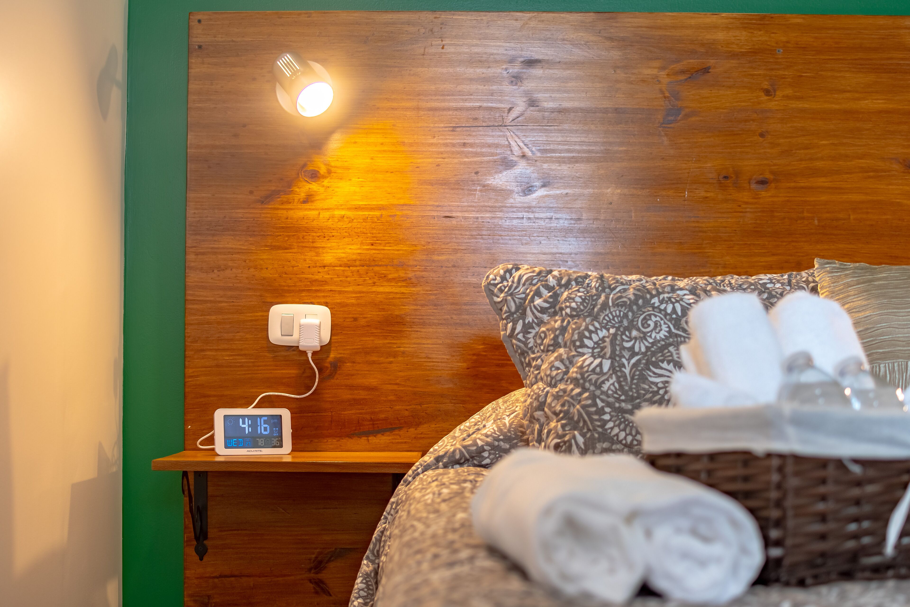 A wooden headboard with a clock and a wall socket, a bed with patterned bedding and rolled towels, and a basket of toiletries.