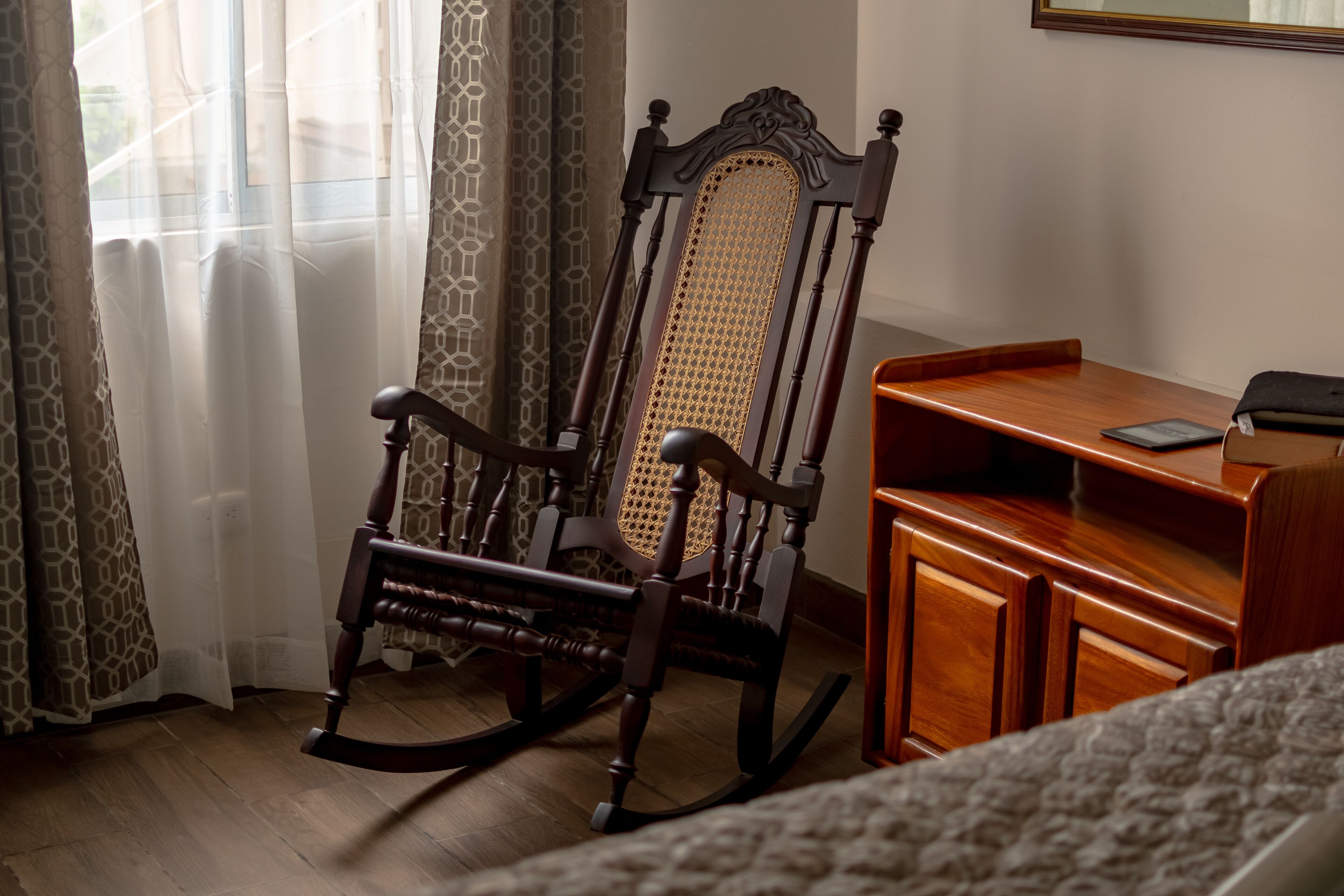 A wooden rocking chair with a woven backrest, a wooden desk with a laptop, and a patterned rug.