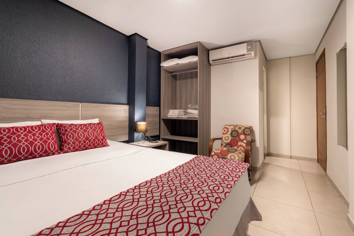 A hotel room with a bed, a wardrobe, a chair, and an air conditioning unit.