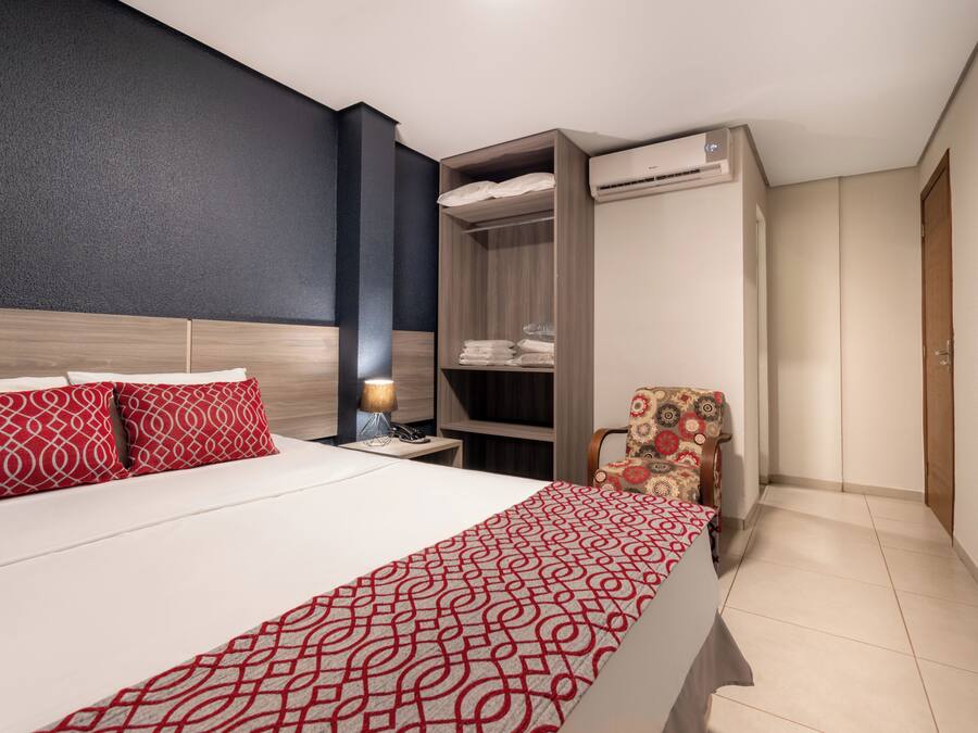 A hotel room with a bed, a wardrobe, a chair, and an air conditioning unit.