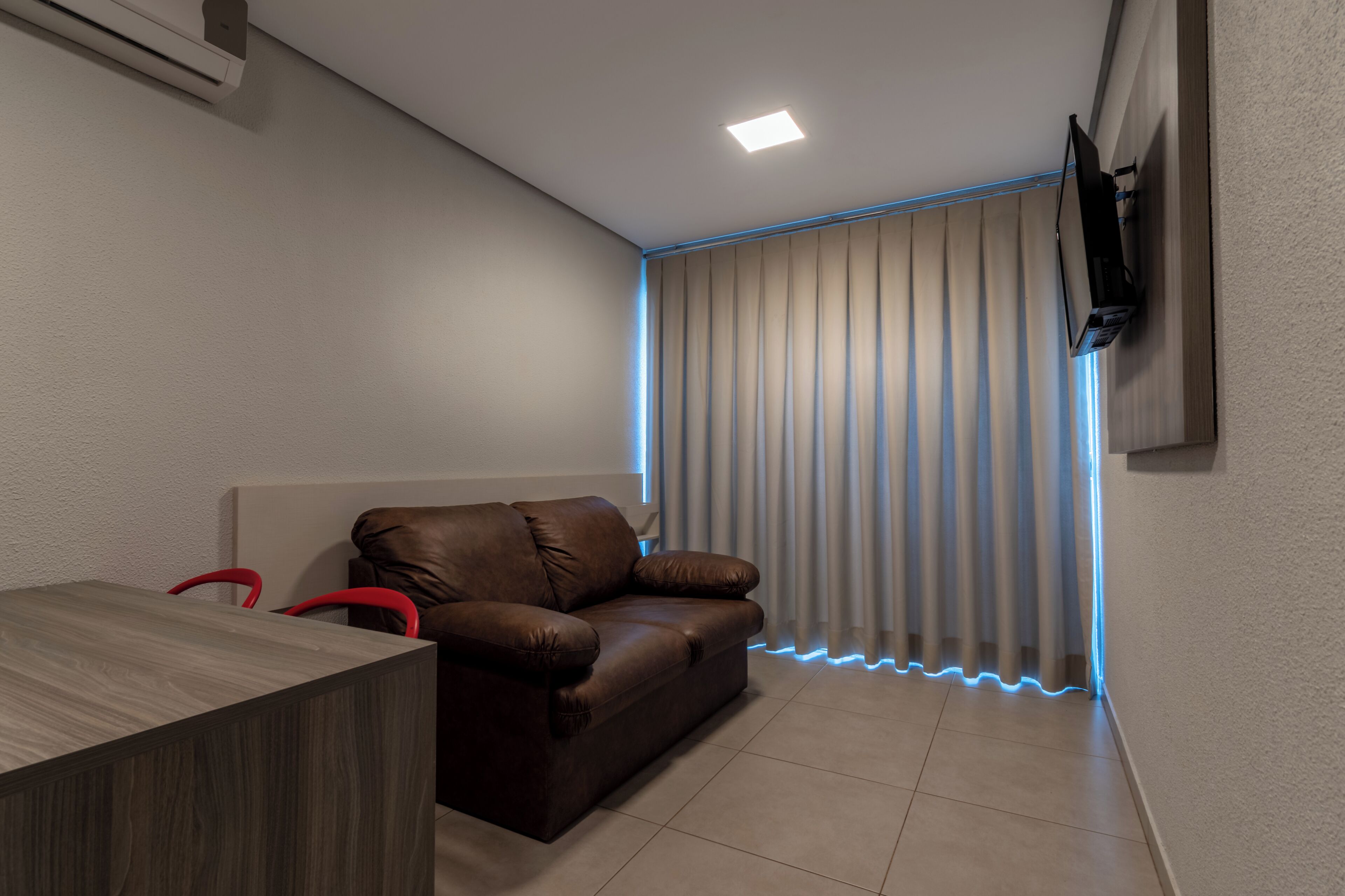 A modern room with a brown leather sofa, a wooden table, a flat-screen TV mounted on the wall, and a window with vertical blinds.