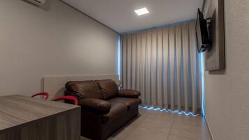 A modern room with a brown leather sofa, a wooden table, a flat-screen TV mounted on the wall, and a window with vertical blinds.