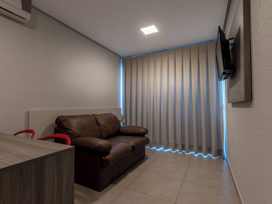 A modern room with a brown leather sofa, a wooden table, a flat-screen TV mounted on the wall, and a window with vertical blinds.