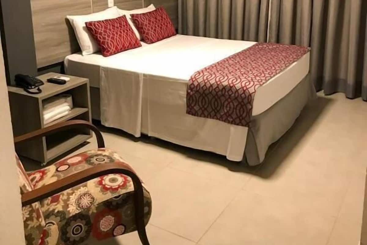 A hotel room with a bed, a nightstand, a chair, and a window with curtains.