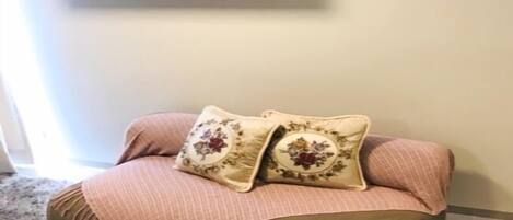 A cozy living room with a sofa, two decorative pillows, a flat-screen TV mounted on the wall, and a fluffy carpet.
