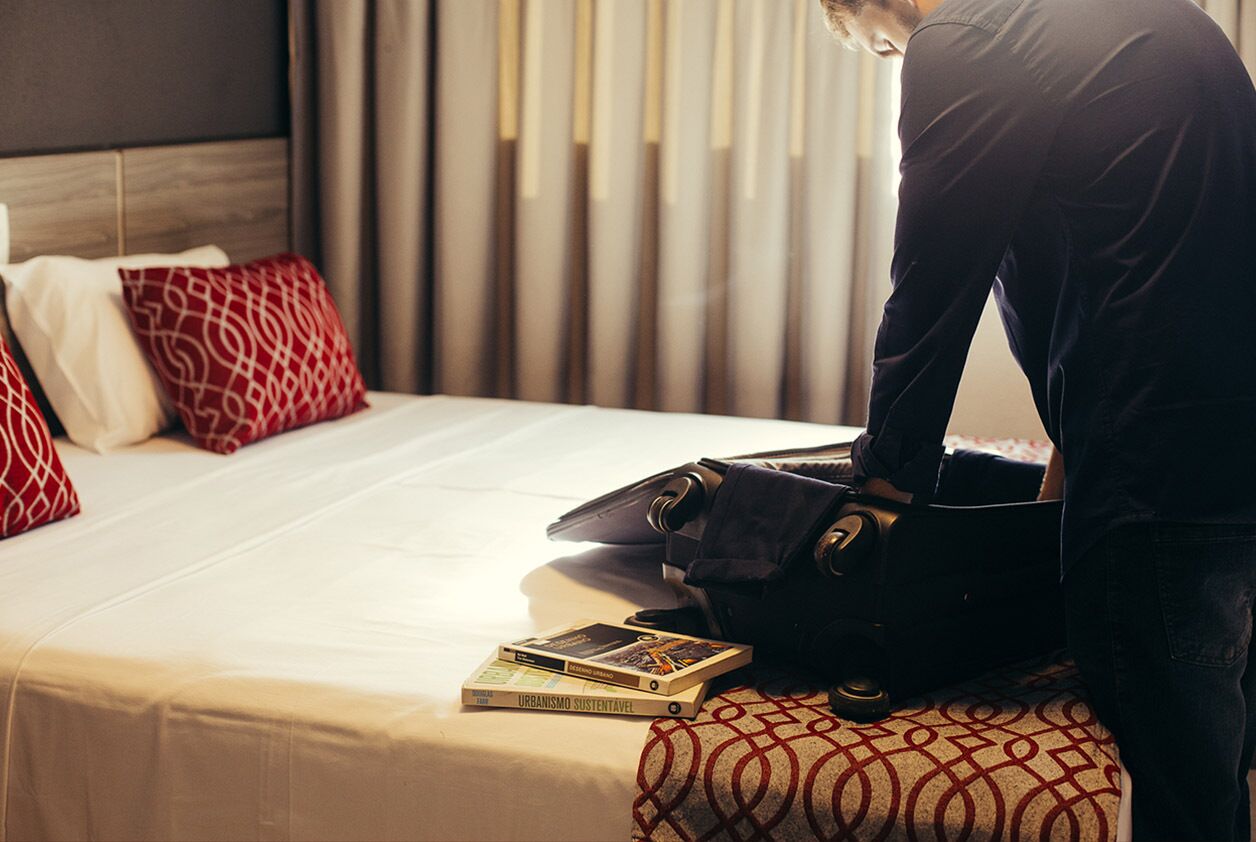 A hotel room with a neatly made bed, a suitcase on the bed, and a book titled "URBANISMO SOSTENTATIVO" on the bed.