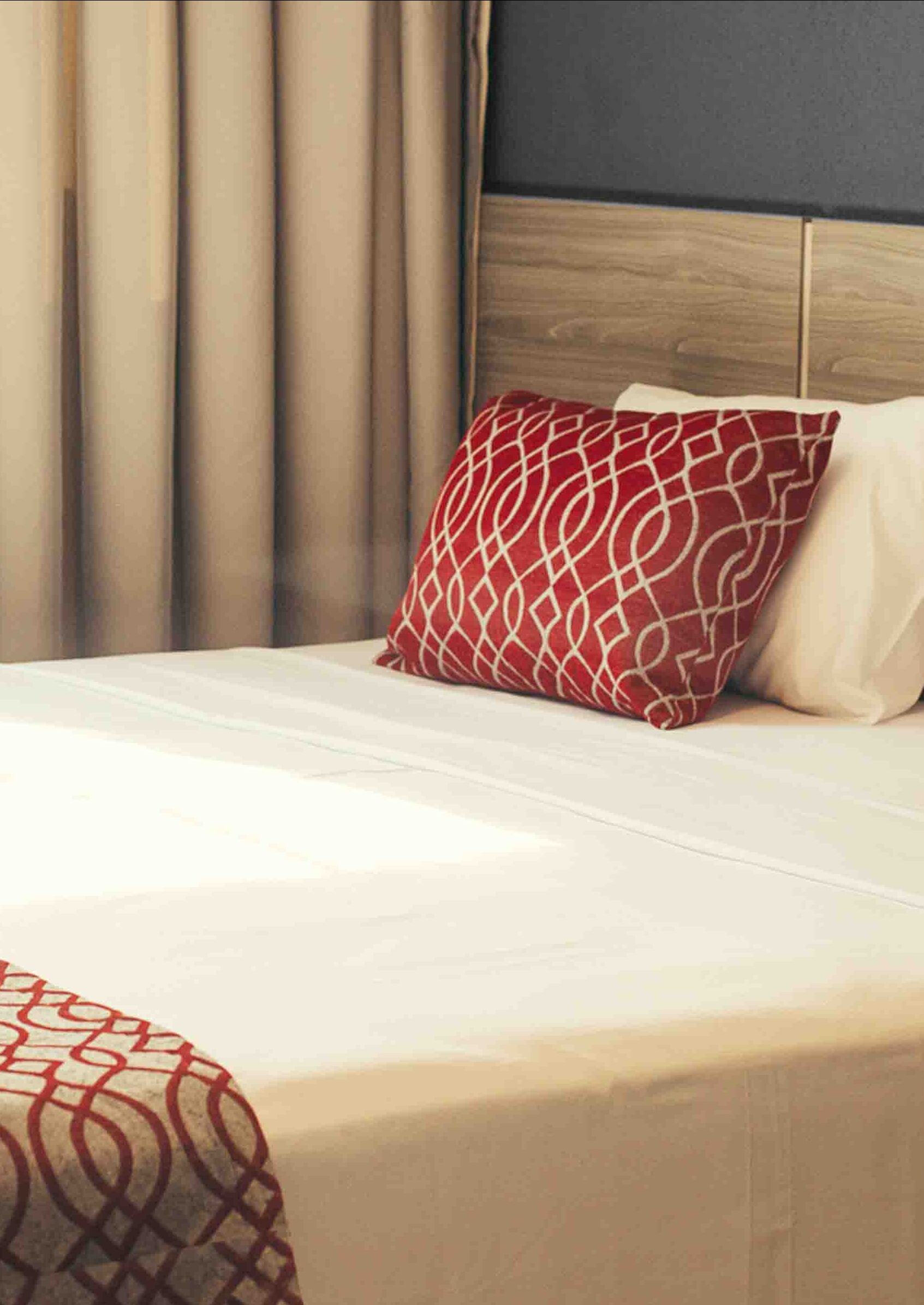 Una cama bien hecha con una colcha estampada, un cojín rojo y blanco, y un cabecero de madera.