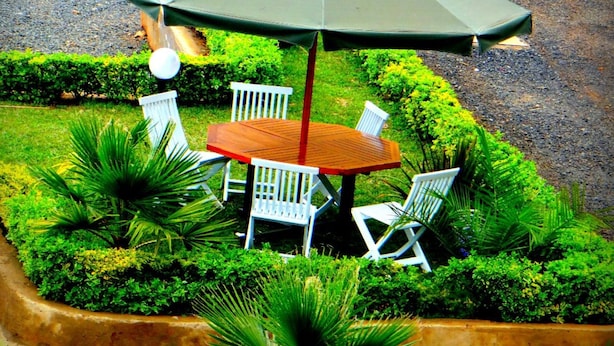 A round wooden table with white chairs under a green umbrella, surrounded by lush greenery and a white fence. - Ridge Apartments (Eldoret)