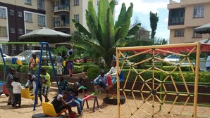 A playground with swings and a climbing structure, surrounded by sand and greenery. - Ridge Apartments (Eldoret)