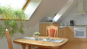 A dining area with a wooden table, chairs, and a potted plant. There is a kitchen in the background with a stove, oven, and cabinets. - Apartment - Apartment Sonnenberg, 25502 (Weener)