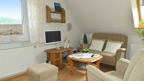 A cozy living room with a sofa, ottoman, wooden coffee table, and a TV. There are potted plants and a clock on the wall. - Apartment - Apartment Sonnenberg, 25502 (Weener)