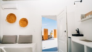 A compact, modern room with a white sofa, two woven baskets on the wall, a small kitchen area, and a view of an outdoor patio. - Mesanto Luxury Suites (Santorini)