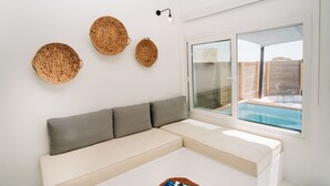 A modern living room with a white sofa, three woven wall decorations, and a large window overlooking a swimming pool. - Mesanto Luxury Suites (Santorini)