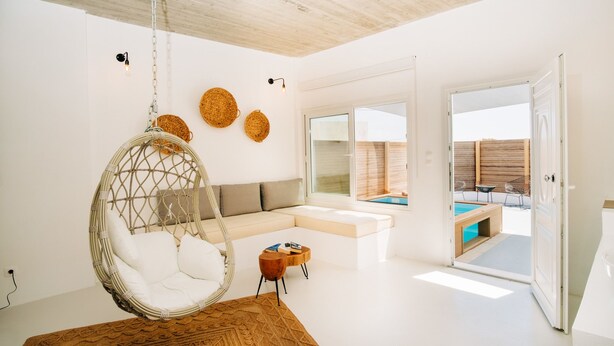 A modern living room with a hanging chair, a sofa, and a view of an outdoor swimming pool. - Mesanto Luxury Suites (Santorini)