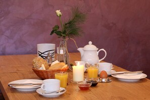 A breakfast setting with a wooden table, a wicker basket with bread, a white pitcher, a white teapot, a lit candle, a white cup, a glass of orange juice, a glass of milk, a small bowl of jam, a plate with a knife and spoon, and a vase with a flower. - Landidyll Landaus Liesetal (Hallenberg)