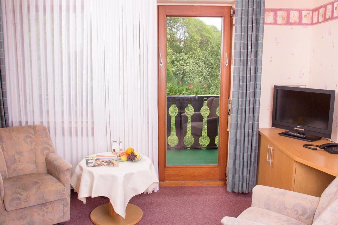 Una habitación pequeña con sofá, una mesa con frutero, televisor y puerta que da a un balcón con barandilla verde.
