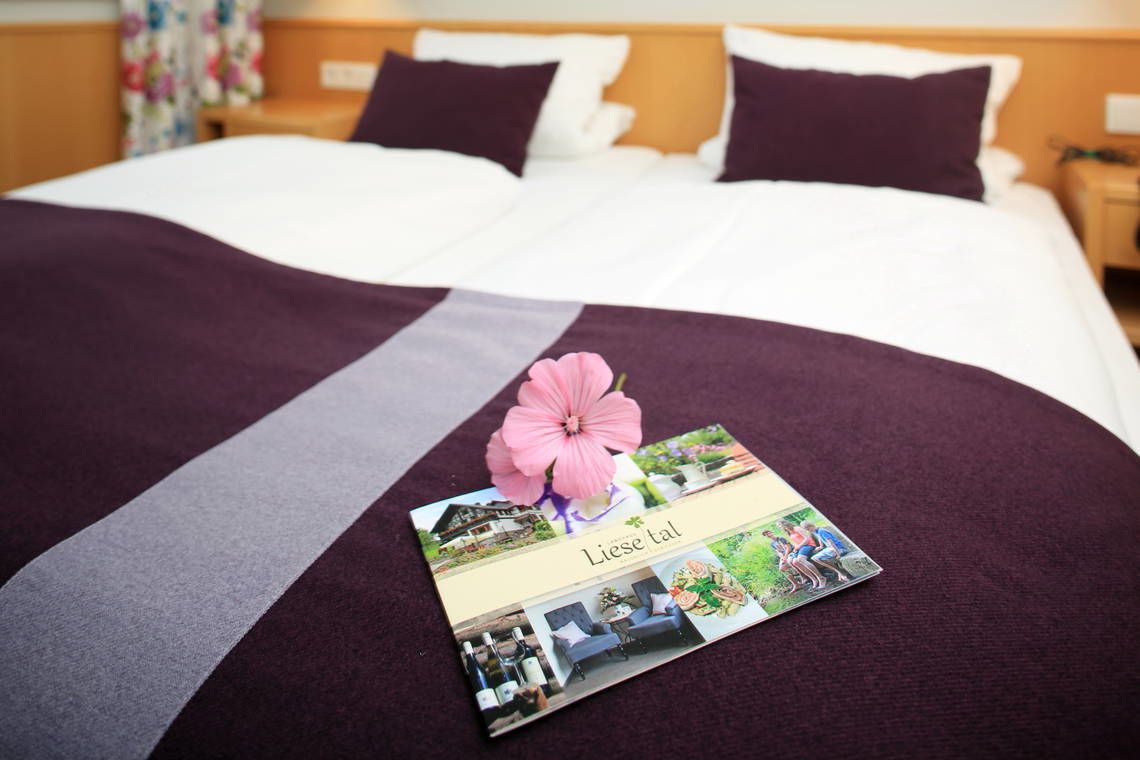A neatly made bed with a purple and white bedspread, a decorative flower, and a brochure titled "Liese tal" on the bed.