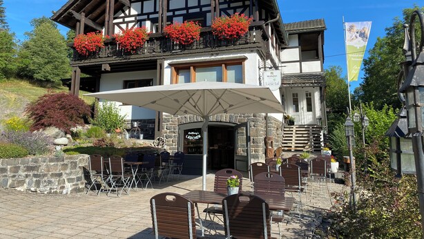 A traditional chalet with a stone facade, wooden balcony with red flowers, outdoor seating, and a sign for 'Lindylow How Restaurant.' - Landidyll Landaus Liesetal (Hallenberg)