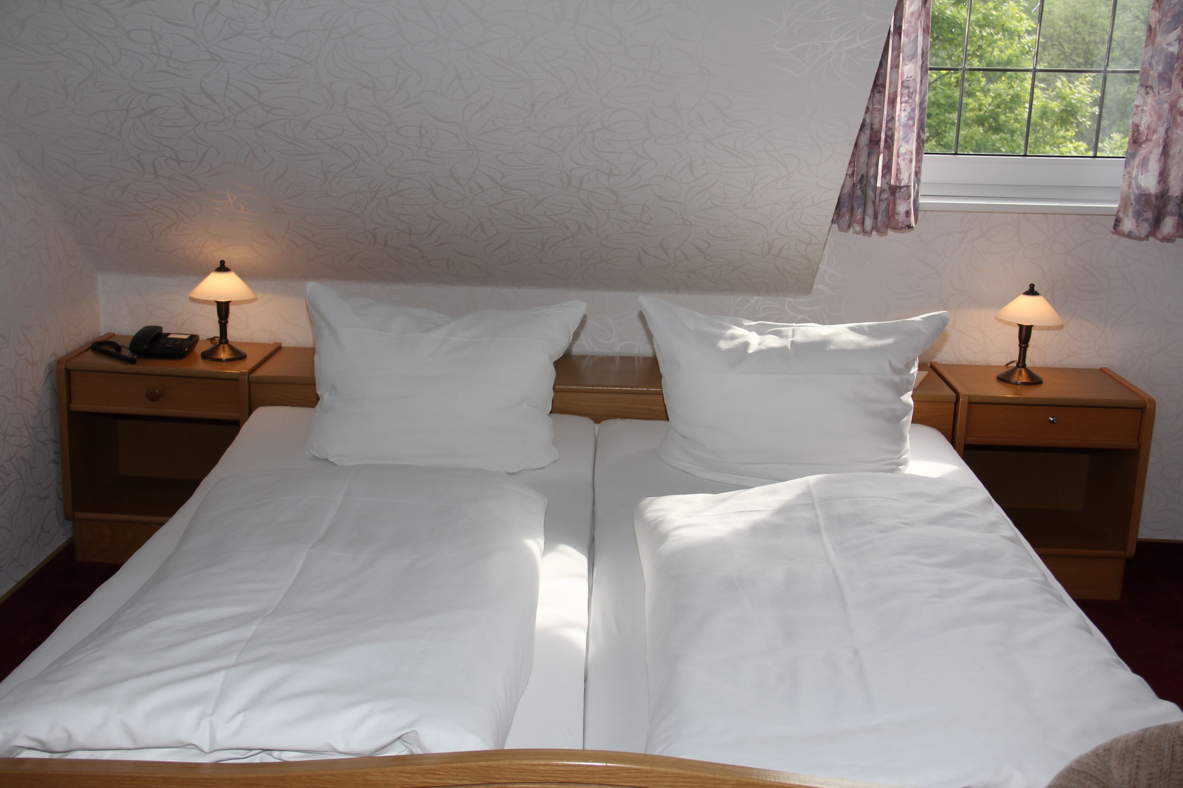 A double bed with white linens, two bedside tables with lamps, a telephone, and a window with curtains.