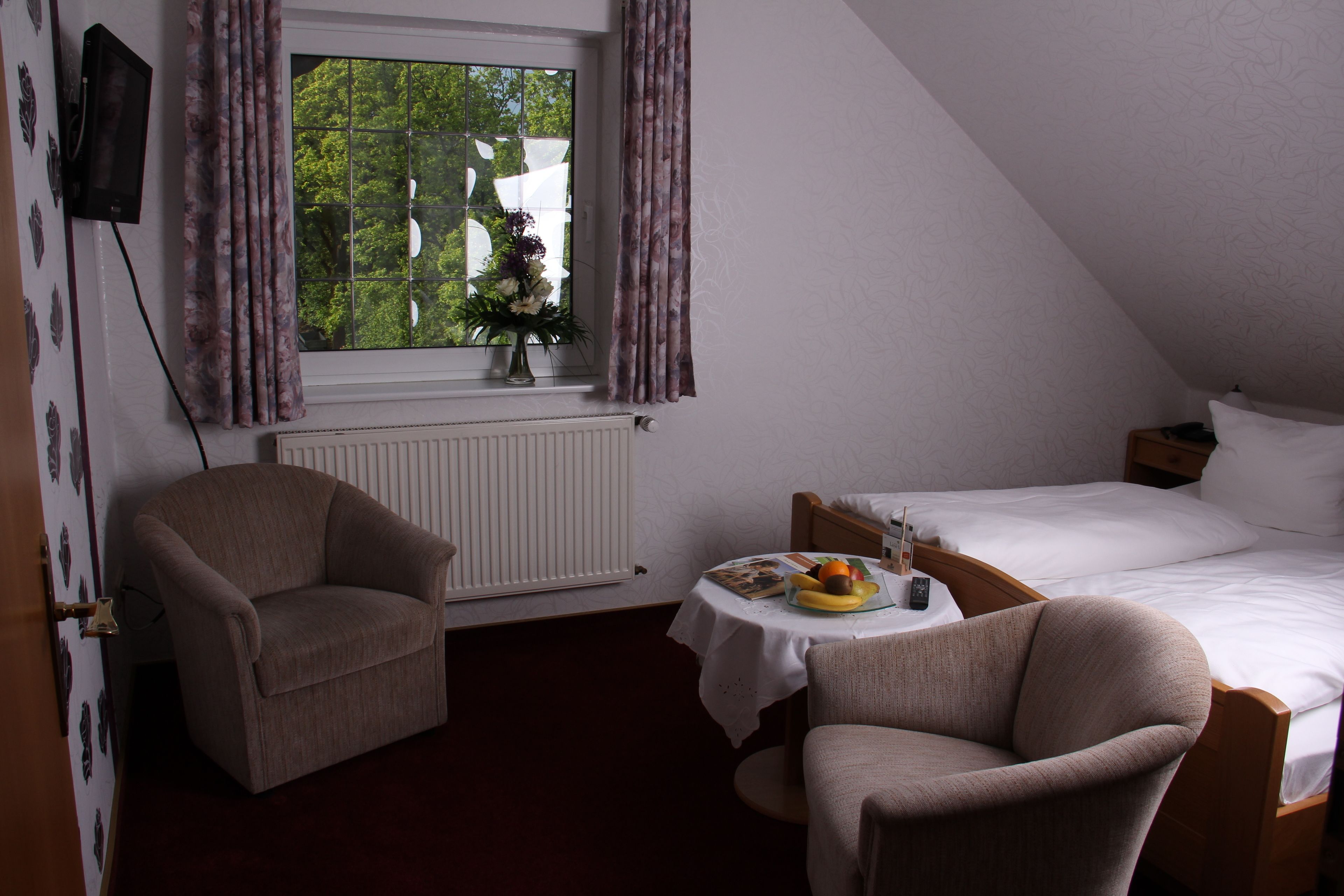 Ein Zimmer mit einem Bett, zwei Sesseln, einem kleinen Tisch mit Früchten, einem Fenster mit Vorhängen und einem Heizkörper.