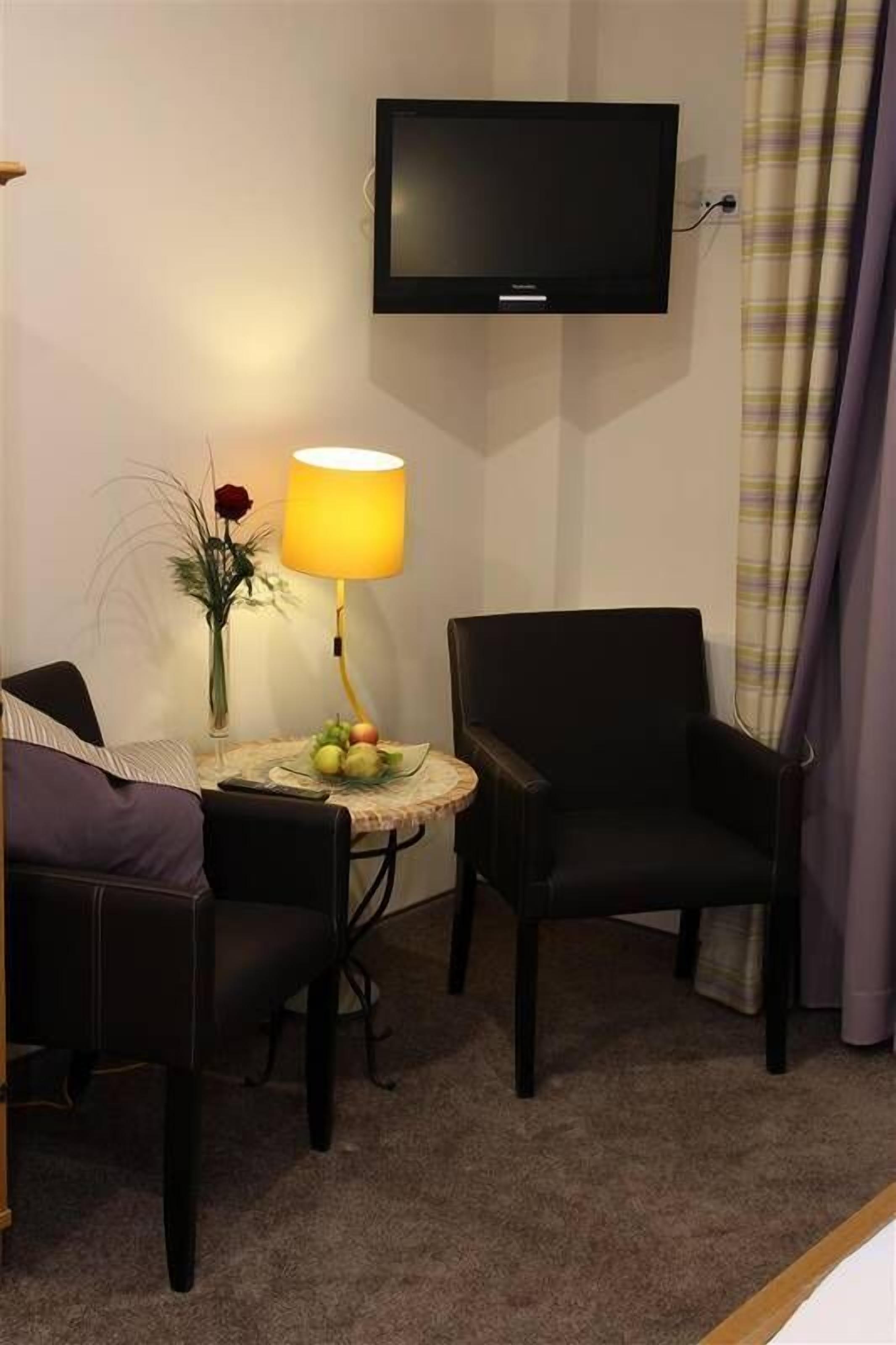 Et hotelværelse med et vægmonteret fladskærms-tv, et lille rundt bord med en lampe, en stol og en vase med blomster.
