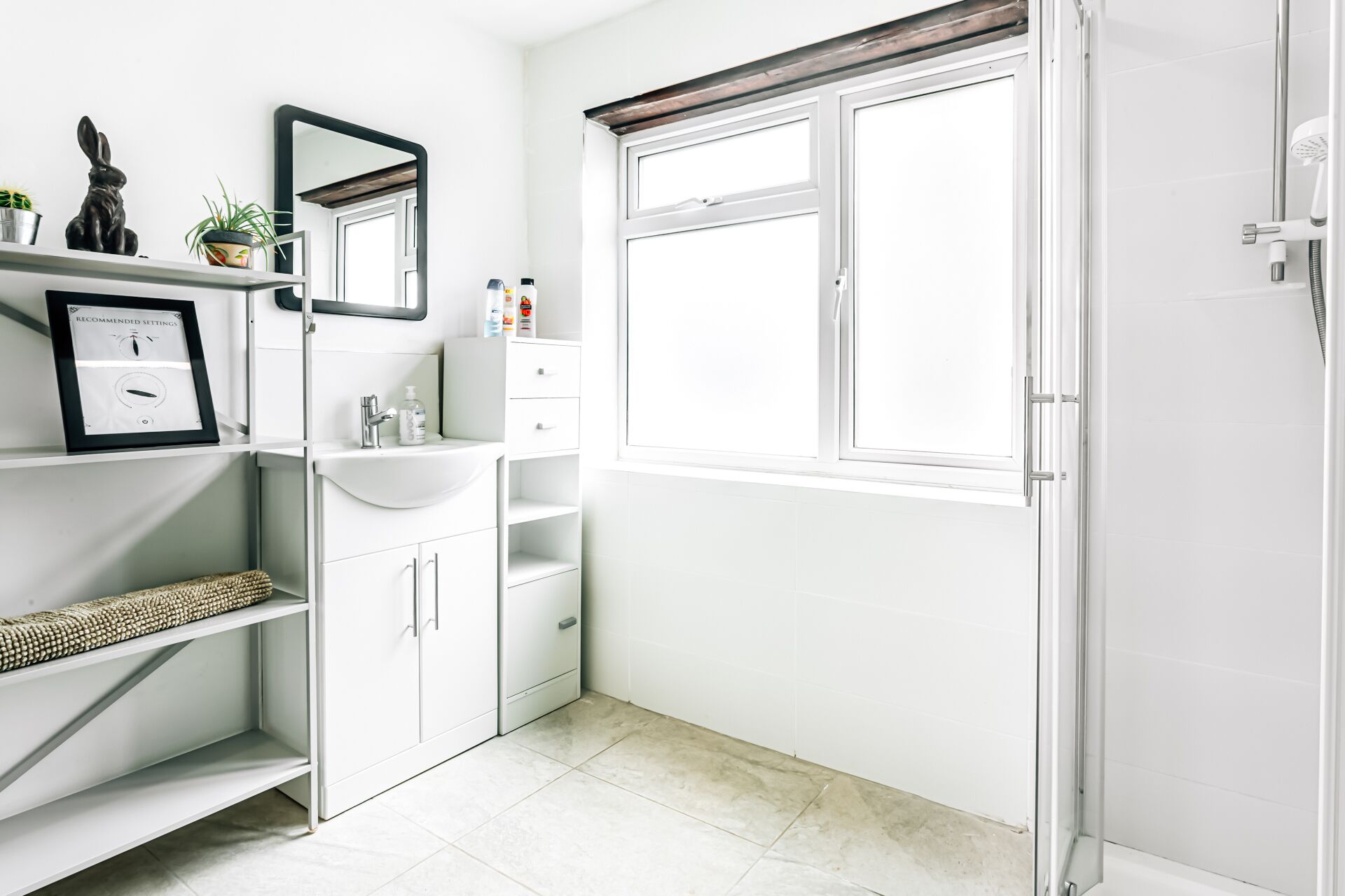 A modern bathroom with a white vanity, a mirror, a window, and a shower area.