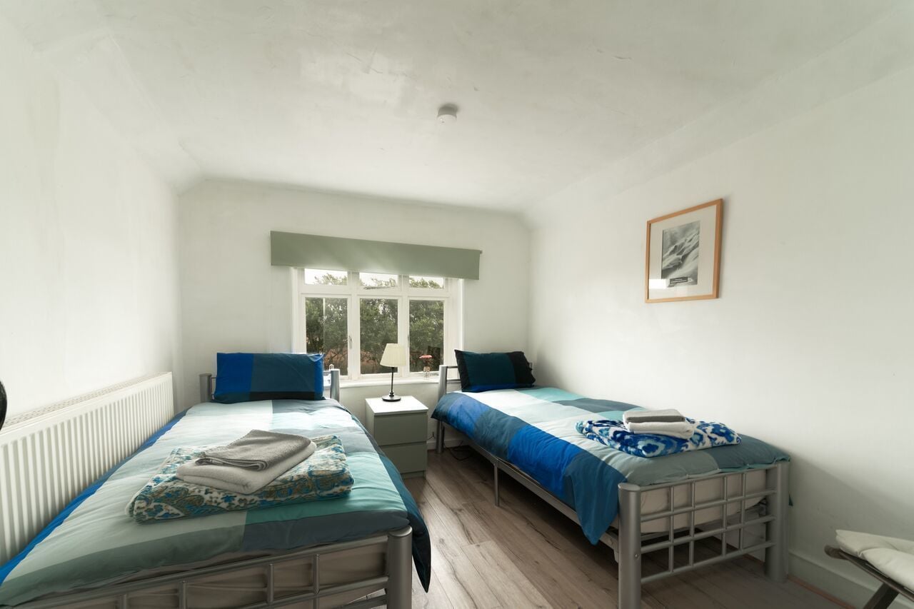 A room with two single beds, a small table, a chair, and a window with a view of trees.