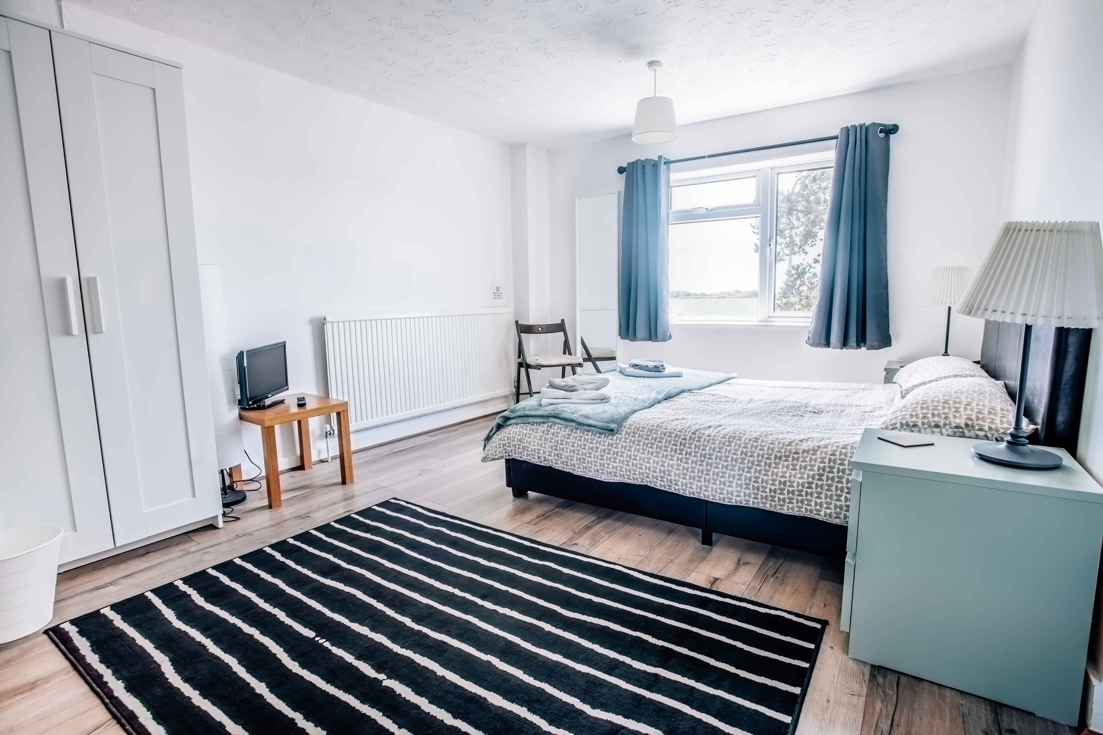Une chambre à coucher comprenant un lit, une table de chevet, une lampe, une chaise, un téléviseur et un radiateur.