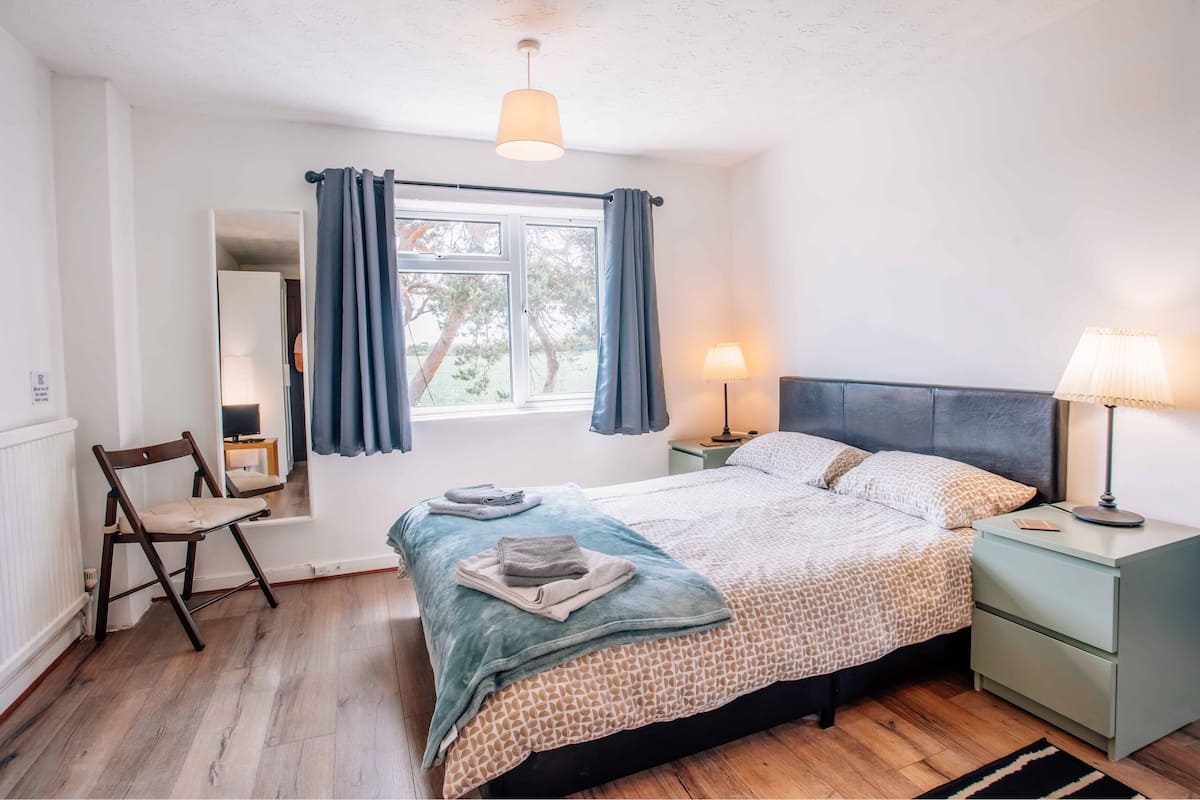 A bedroom with a bed, two bedside tables with lamps, a chair, a window with curtains, and a view of trees outside.