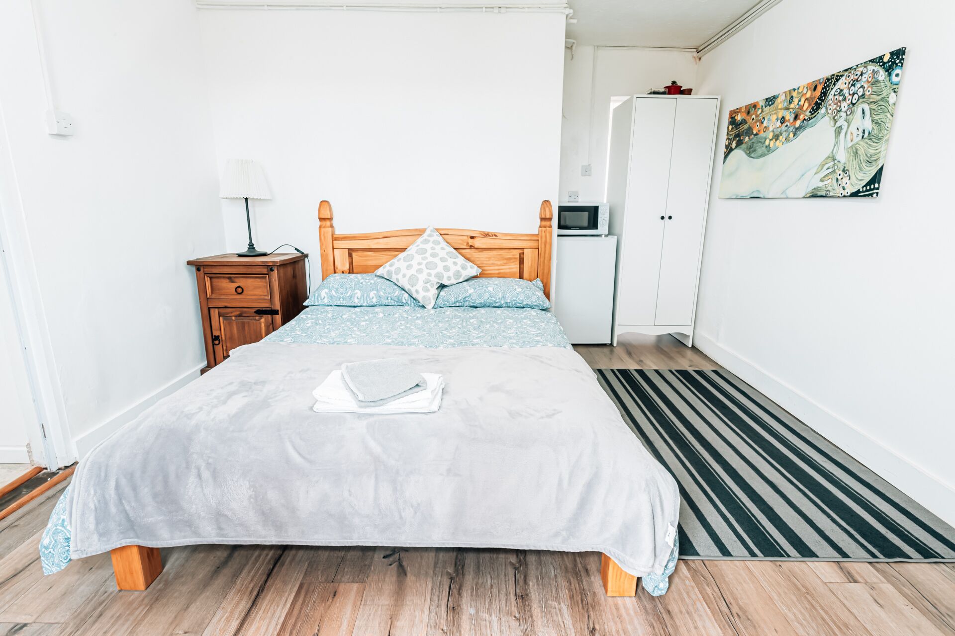 Un dormitorio ordenado con cama, mesita de noche, lámpara, armario y una obra de arte en la pared.