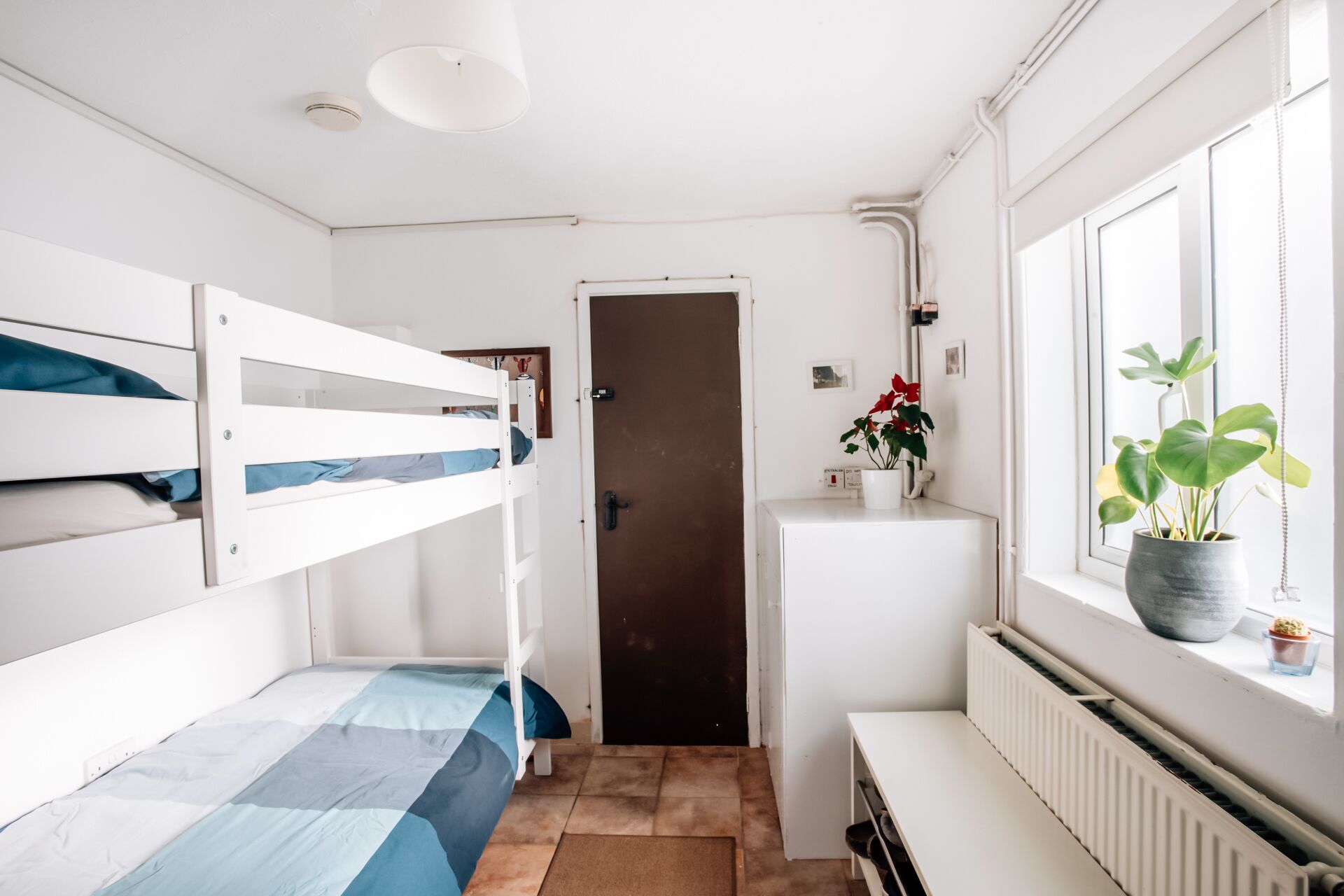 A small, compact room with bunk beds, a radiator, a window with a plant, and a door leading to another room.