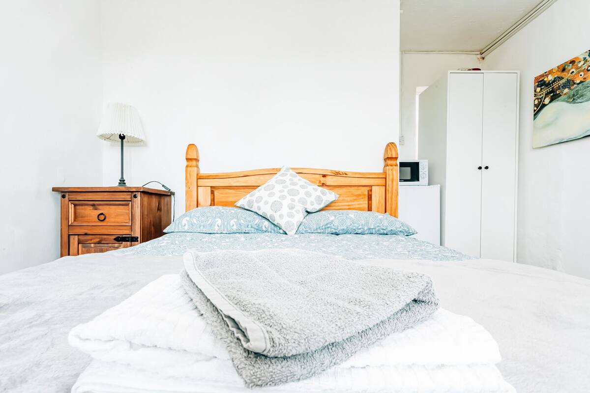 A neatly made bed with a grey blanket and patterned pillows, a wooden nightstand with a lamp, and a white wardrobe in a simple room with white walls.