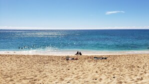 Ein sandiger Strand mit klarem, blauem Meer, wenigen Personen und Strandausstattung.