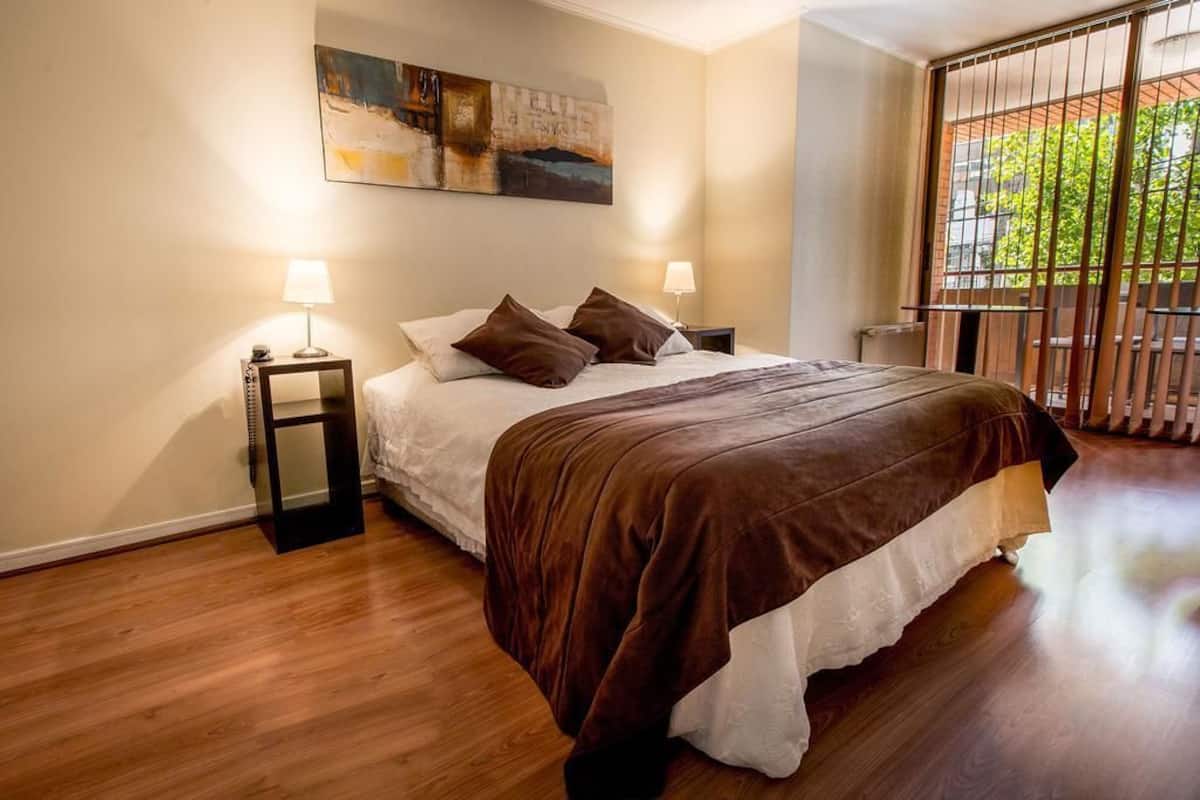 Une chambre avec un grand lit, deux tables de chevet avec des lampes, un tableau au mur et une vue par une fenêtre ornée d’une rambarde en métal.