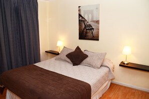 A neatly made bed with a brown blanket and pillows, two bedside tables with lamps, a wall-mounted picture of a bridge, and a wooden floor.