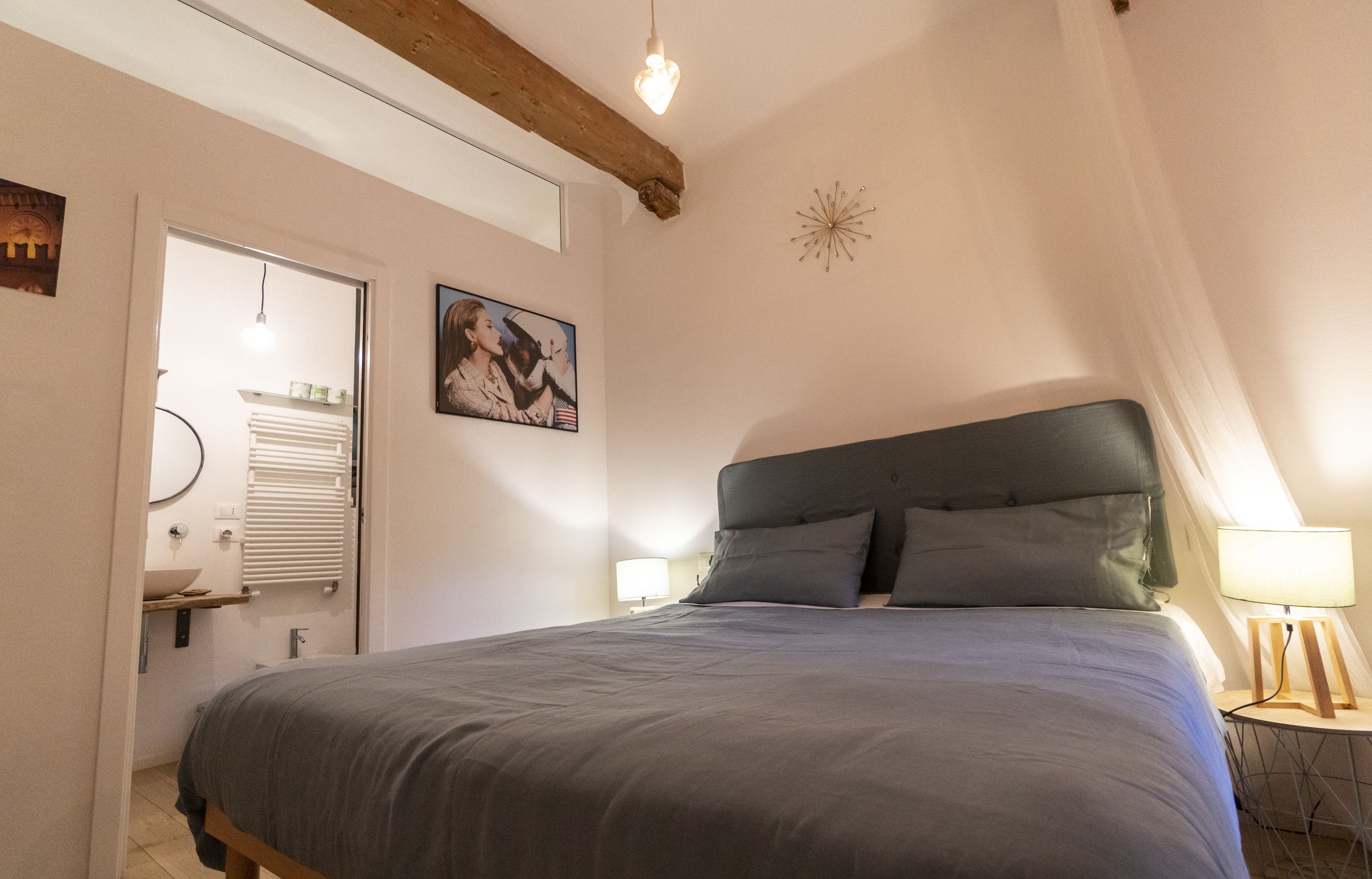 A bedroom with a bed, a bedside table with a lamp, a wall-mounted picture, and a window with blinds.