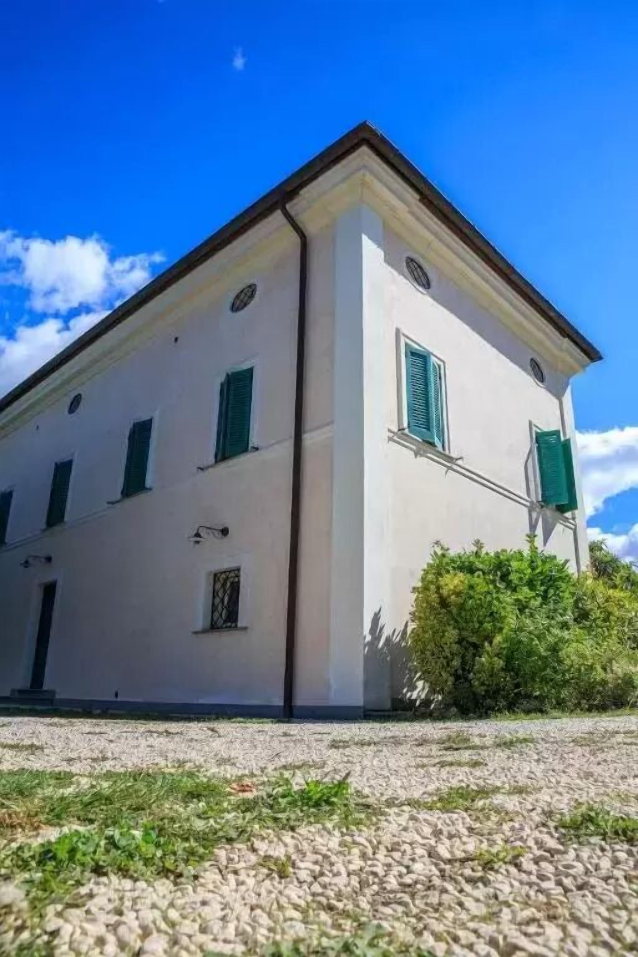 A two-story building with a white facade and green shutters, a gravel driveway, and a clear blue sky.