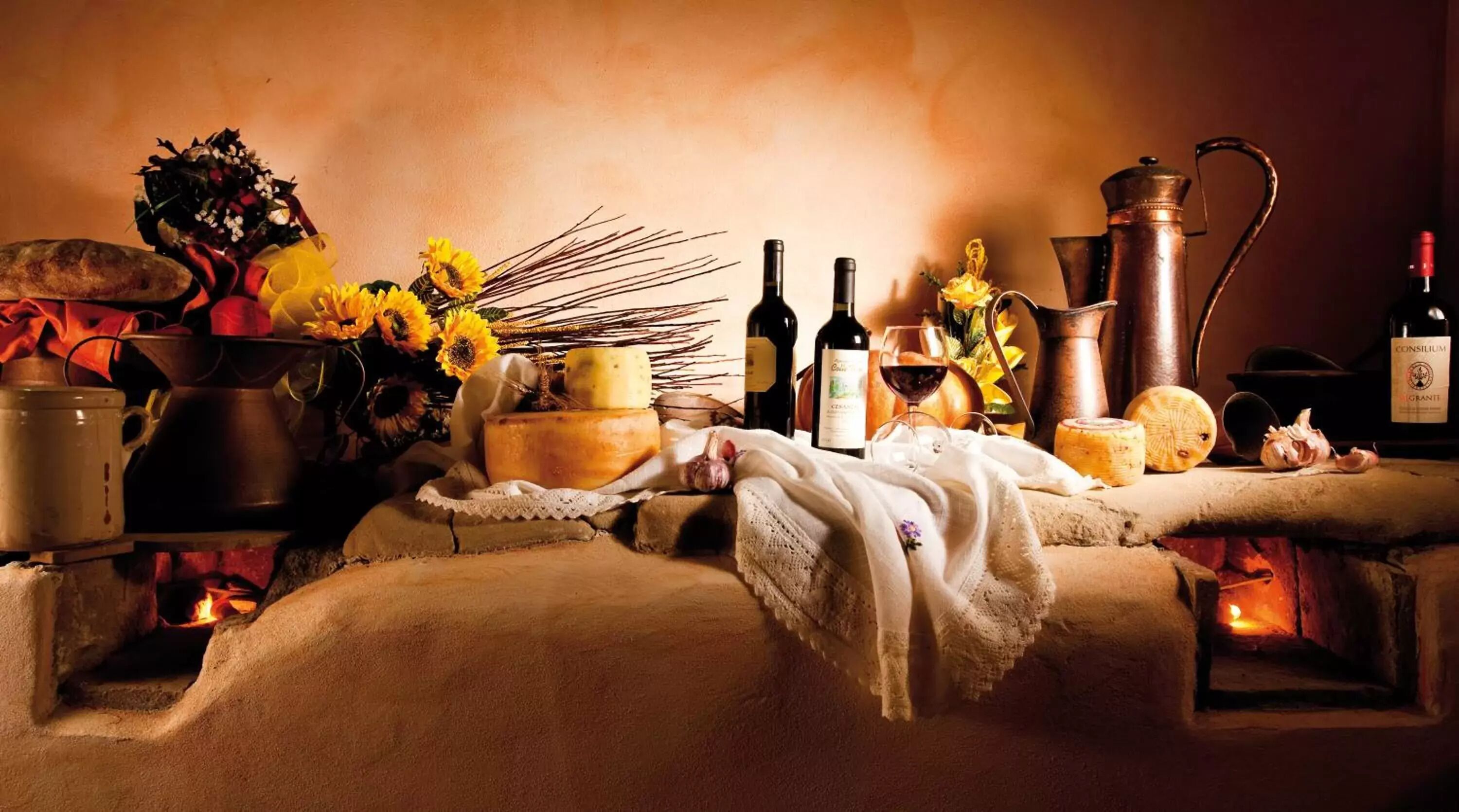 Un ambiente de cocina rústica con fuego, utensilios de cocina, un queso redondo, botellas de vino y un ramo de girasoles.