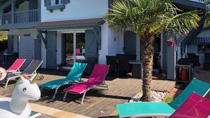 A two-story house with a covered patio area featuring lounge chairs and a palm tree. - Villa Andernos (Andernos-les-Bains)