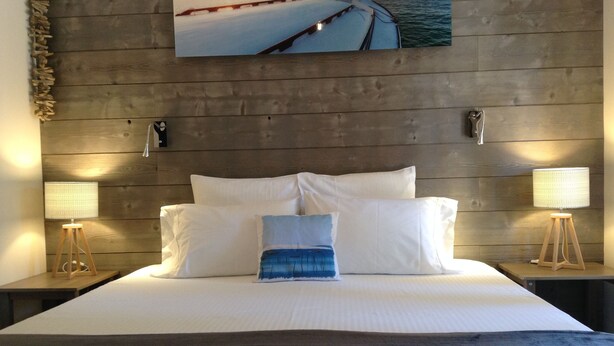A hotel room with a wooden headboard, a large bed with white linens, two bedside lamps, and a painting of a sunset over water. - Villa Andernos (Andernos-les-Bains)